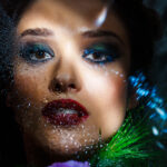 Close up fashion portrait of young beautiful brunette girl with bright make up looking at camera through misted glass, lying among white and purple eustomas.