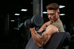 Close up of handsome bodybuilder training biceps with dumbbell on bench. Close up of muscular sportsman with perfect body posing in gym in dark atmosphere. Concept of bodybuilding.
